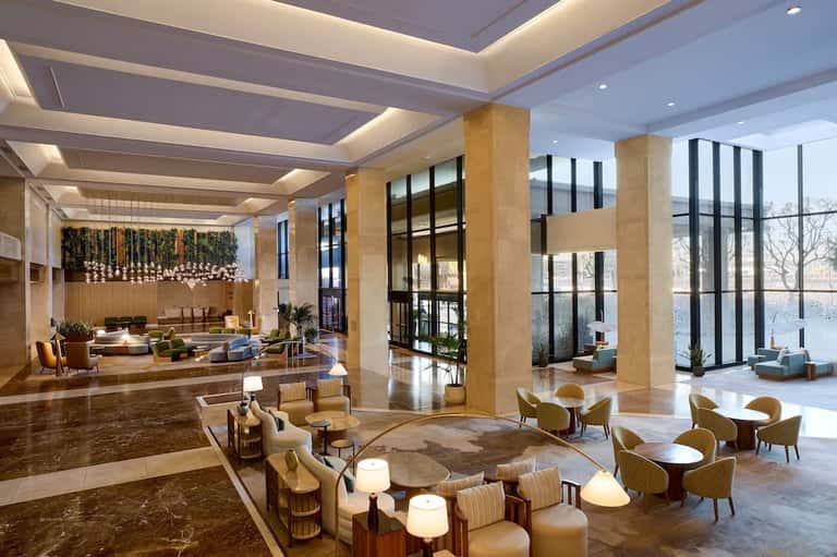 Luxury hotel lobby with tall columns, modern lighting, seating areas, and floor-to-ceiling windows overlooking water