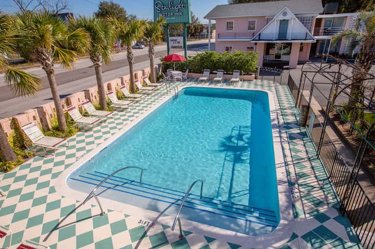 Retro motor inn courtyard with turquoise pool, checkered pavement, pink buildings, palm trees, and vintage signage