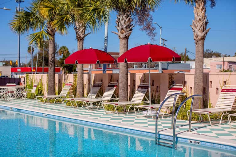 Poolside area with red umbrellas, lounge chairs, palm trees, pink building, and clear blue pool water