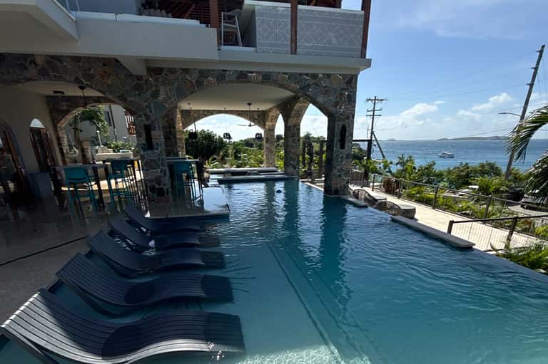 Infinity pool under stone arches with ocean views and lounge chairs on wooden deck