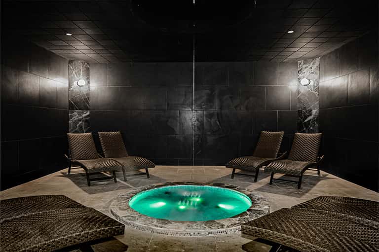 Luxurious spa area with glowing turquoise hot tub, marble walls, and woven lounge chairs