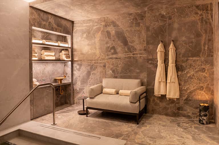 Luxury spa relaxation room with marble walls, beige lounge seating, and hanging robes with subtle lighting