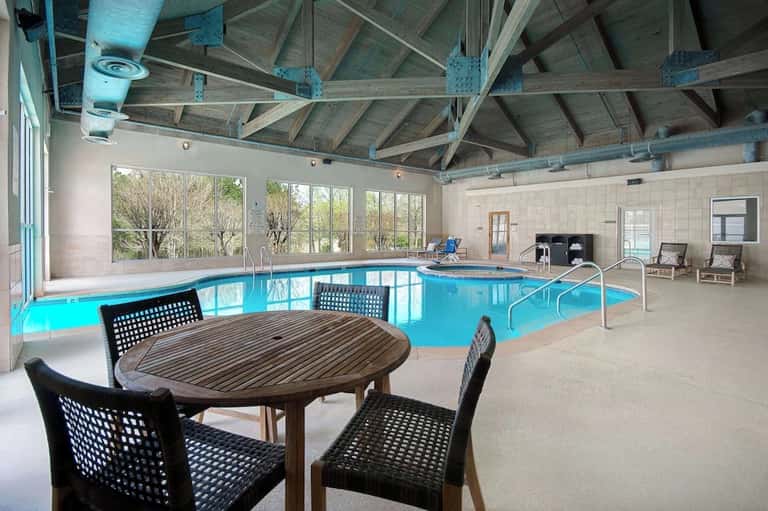 Indoor heated pool facility with bright blue water, wooden table, exposed ceiling beams, and large windows