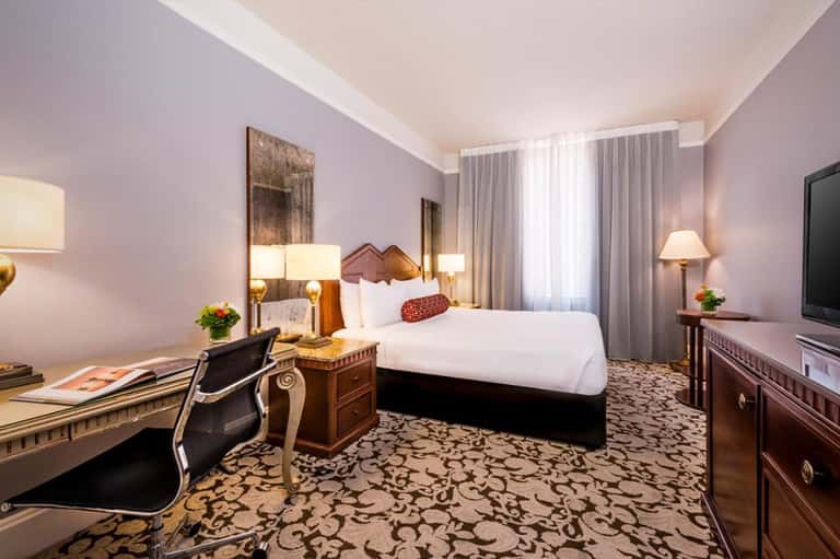 Elegant hotel bedroom with purple walls, four-poster bed, desk area, and patterned carpet