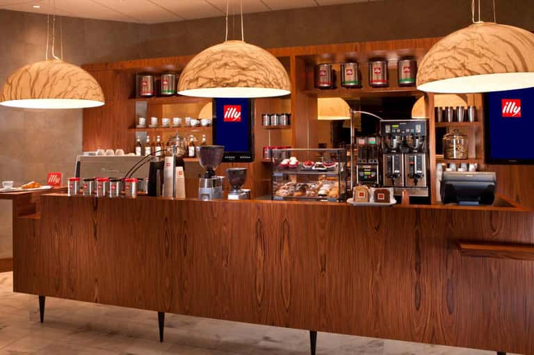 Modern coffee shop counter with wooden cabinetry, hanging dome lights, and Illy coffee branding
