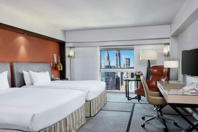 Upscale hotel room with twin beds, orange accent wall, city skyline view, and modern furnishings