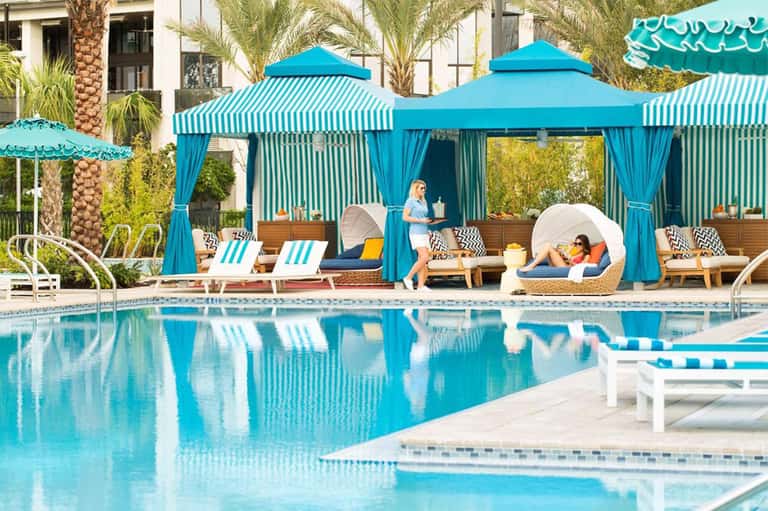Resort pool deck with turquoise striped cabanas, loungers, palm trees, and guests relaxing poolside