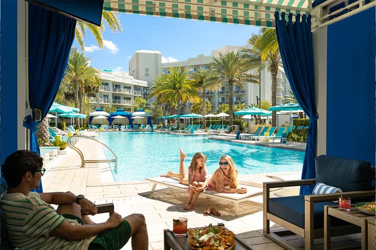 Family relaxing poolside under pergola with view of resort pool and palm trees