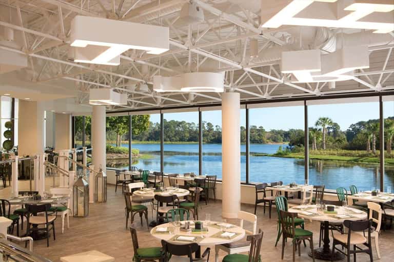 Modern restaurant interior with floor-to-ceiling windows overlooking river, white ceiling beams, and dining tables