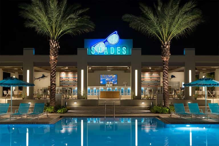 Modern poolside bar at night with neon 'SHADES' sign and blue umbrellas