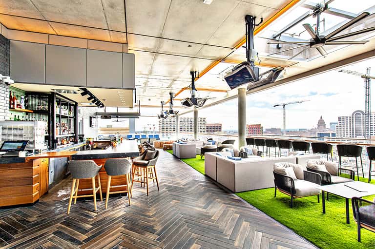 Modern rooftop lounge with bar, seating areas, green turf, and city skyline views including a capitol building