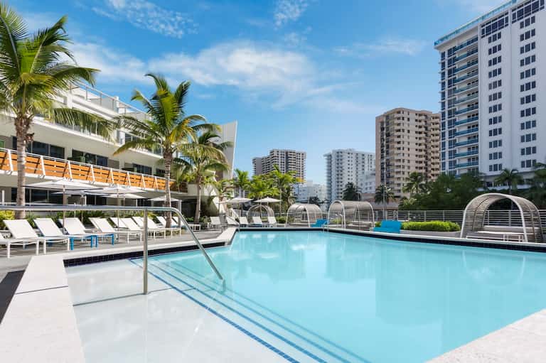 Resort pool deck with palm trees, white loungers, parasols, and multi-story residential buildings in background