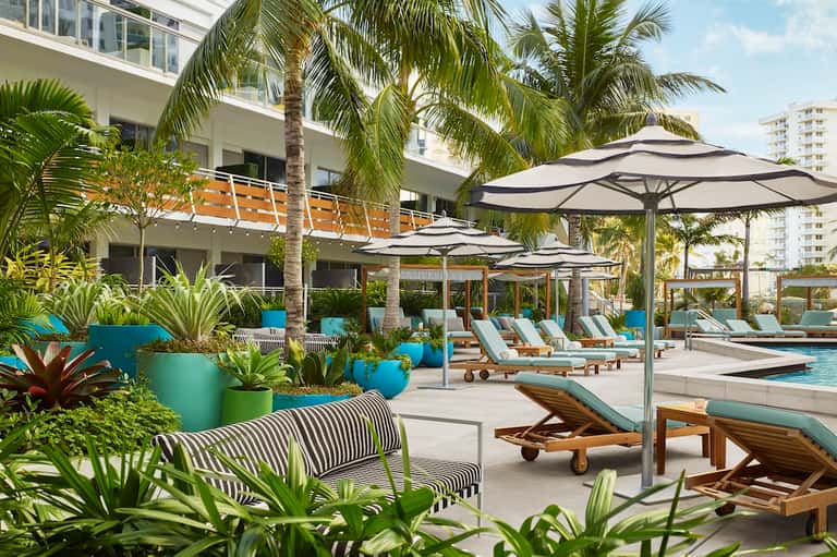 Modern poolside lounge area with gray umbrellas, wooden loungers, and palm trees