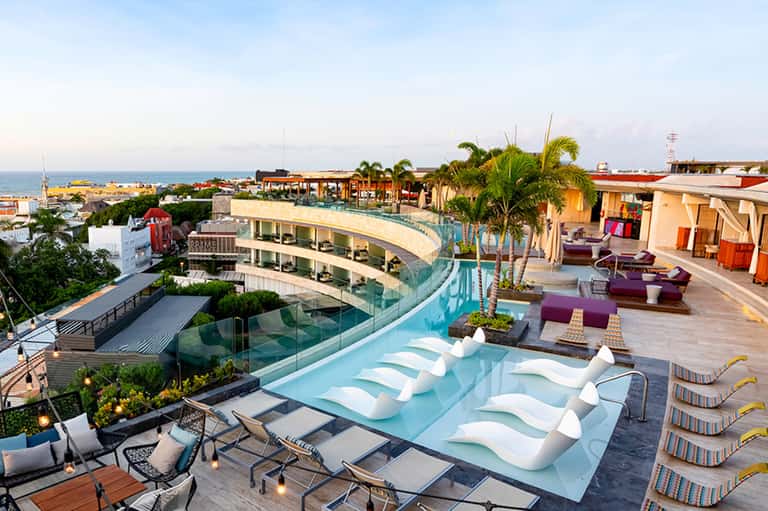 Rooftop infinity pool at sunset with ocean views, lounge chairs, palm trees, and coastal resort architecture