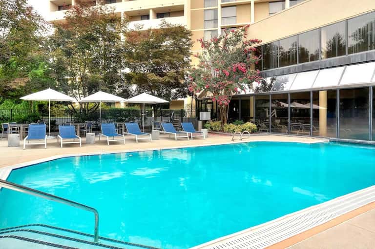 Resort pool surrounded by blue lounge chairs and white umbrellas with modern building and flowering trees in background