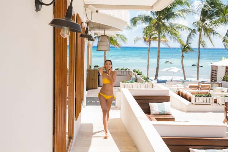 Beachfront lounge area with white columns, palm trees, ocean view, and woman in yellow bikini