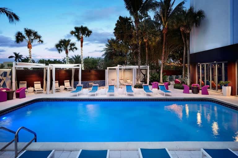 Resort pool at twilight with palm trees, modern cabanas, colorful seating, and building facade