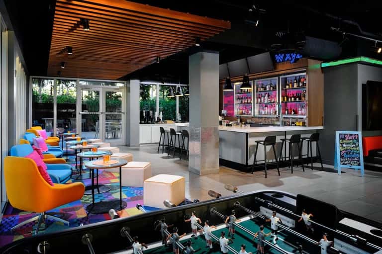 Vibrant hotel lounge featuring colorful chairs, bar with neon sign, foosball table, and outdoor patio