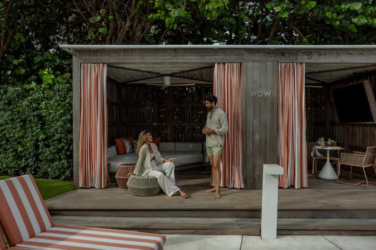 Outdoor resort cabana with striped curtains, lounge seating, and couple relaxing under mature trees.