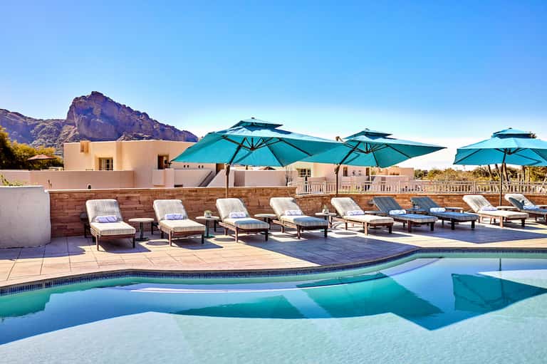 Luxury resort pool with turquoise umbrellas, white lounge chairs, mountain backdrop, and modern architecture