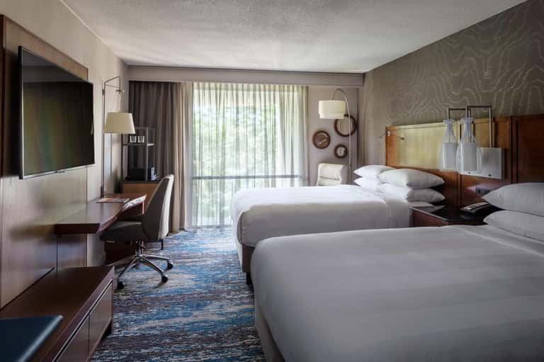 Modern hotel room with two queen beds, desk, TV, and sliding glass doors to balcony