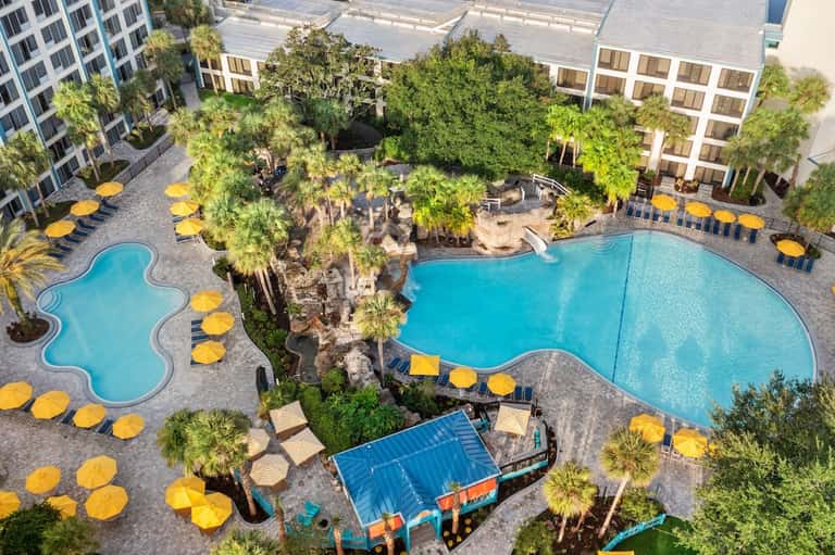 Aerial view of resort pool complex with yellow umbrellas, palm trees, and surrounding buildings