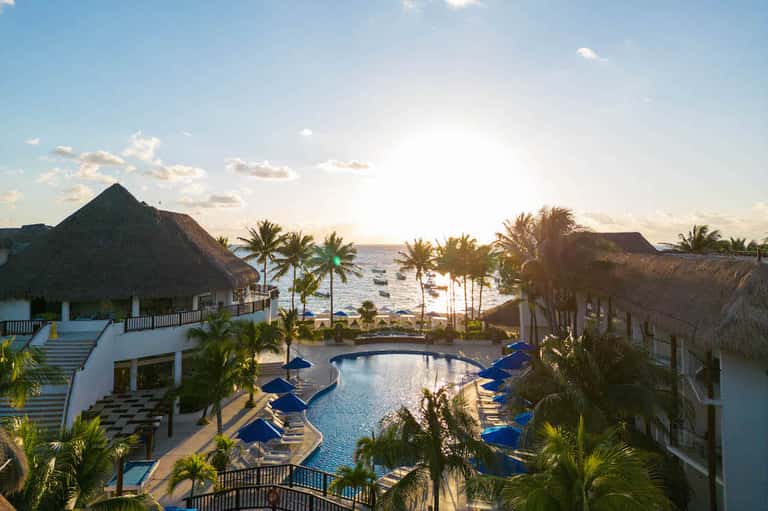 Beachfront resort pool with palm trees, tiki-roofed structures, and ocean view at golden hour