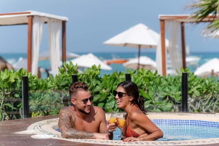 Couple enjoying drinks in infinity pool overlooking ocean with white cabanas and beach umbrellas