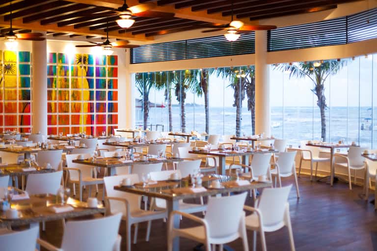 Upscale dining restaurant with colorful artwork wall, white chairs, ocean views, and palm tree columns