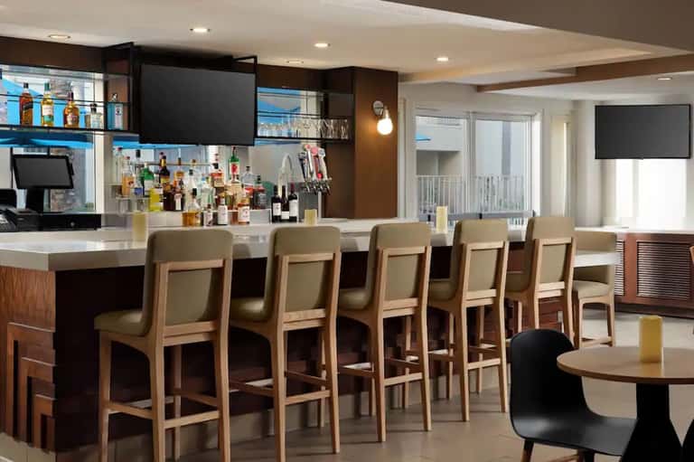 Modern hotel bar with wood accents, barstool seating, full liquor display, and flat-screen TV