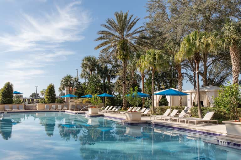 Resort pool surrounded by palm trees, blue umbrellas, lounge chairs, and white cabanas