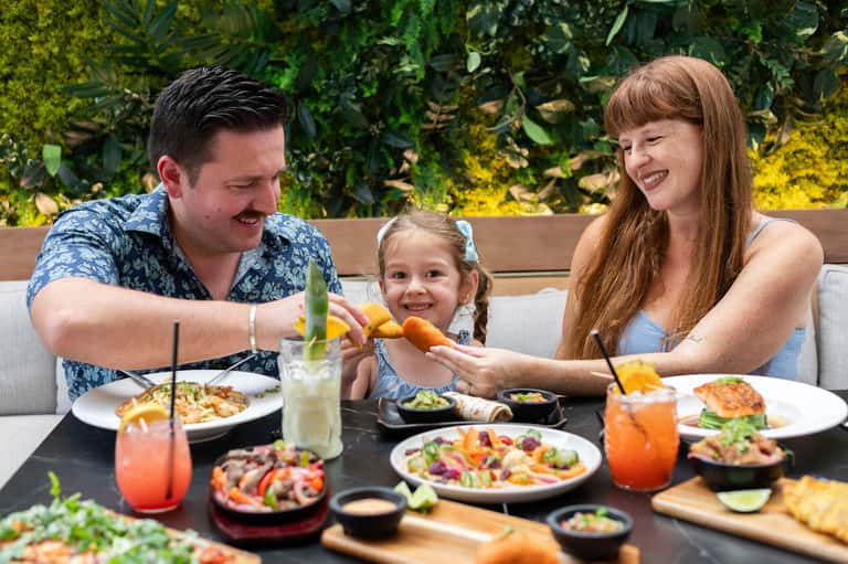 Family dining outdoors with colorful appetizer plates, tropical cocktails, and living plant wall backdrop