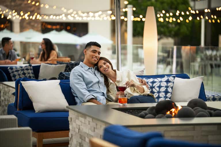 Couple enjoying drinks by outdoor fireplace with string lights and comfortable seating