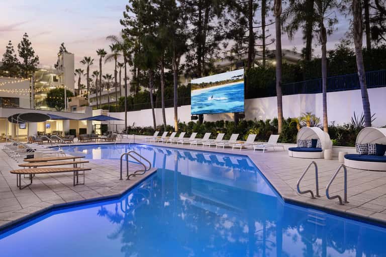 Resort pool at dusk with lounge chairs, palm trees, white cabanas, and lit buildings