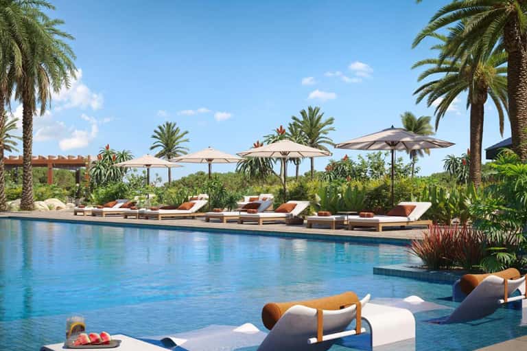 Resort pool with lounge chairs, umbrellas, palm trees, and tropical landscaping under clear blue sky