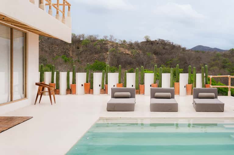 Modern patio with infinity pool, three gray lounge chairs, potted cacti, mountain views, minimalist white design