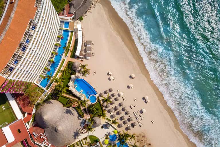 Aerial view of beachfront resort with pools, palm trees, and sandy beach meeting turquoise ocean