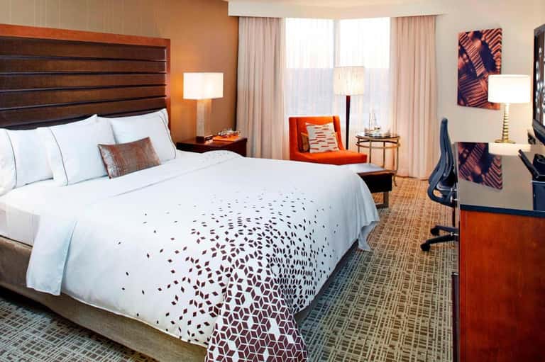 Hotel bedroom with warm orange walls, wooden headboard, white bedding, and sitting area with window views