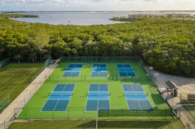 Aerial view of multiple blue tennis courts on manicured grass with waterfront and trees in background