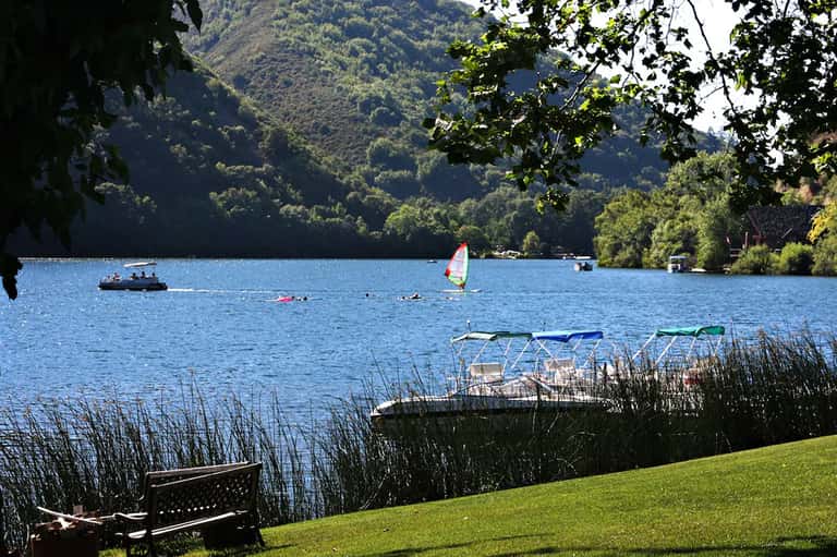 Scenic lake view with sailboat, forested mountains, and wooden bench on grassy shore