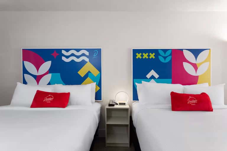 Modern hotel room with two queen beds, colorful geometric headboard artwork, and red accent pillows