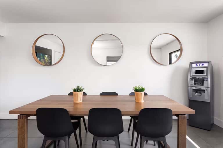 Minimalist meeting room with wooden table, black chairs, three round mirrors, and ATM machine on white walls.