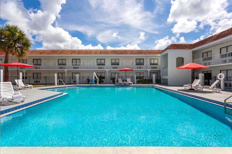 Outdoor pool surrounded by white hotel buildings, lounge chairs, and red umbrellas under clear sky