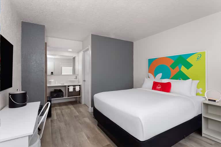 Modern hotel room with queen bed, ensuite bathroom, desk, and colorful abstract headboard art