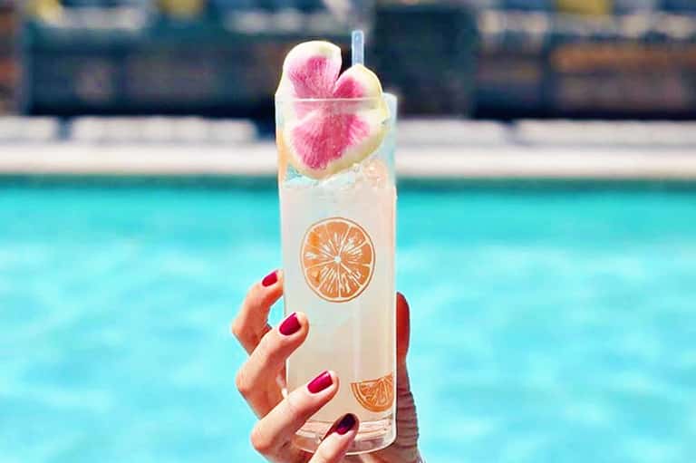 Tall clear cocktail with citrus slices and pink flower garnish held over pool background