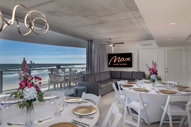 Modern beachfront event space with ocean views, dark sectional seating, elegant dining setup, and decorative flowers