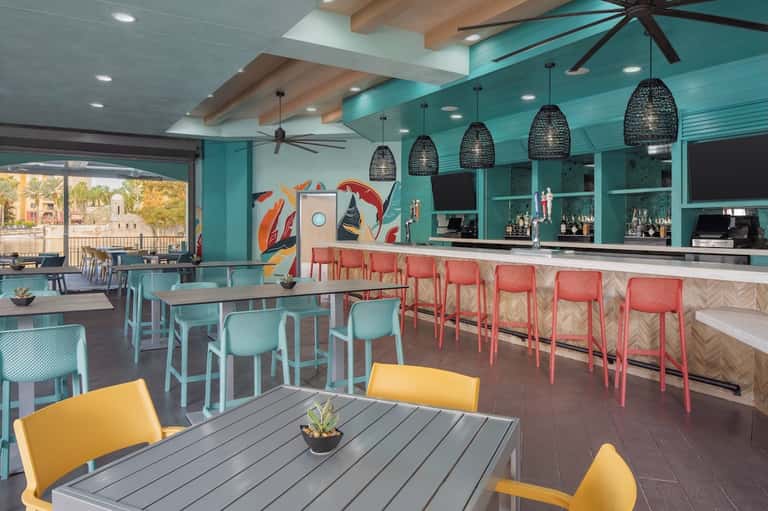 Colorful resort bar and dining area with turquoise walls, coral chairs, yellow accents, and ocean view