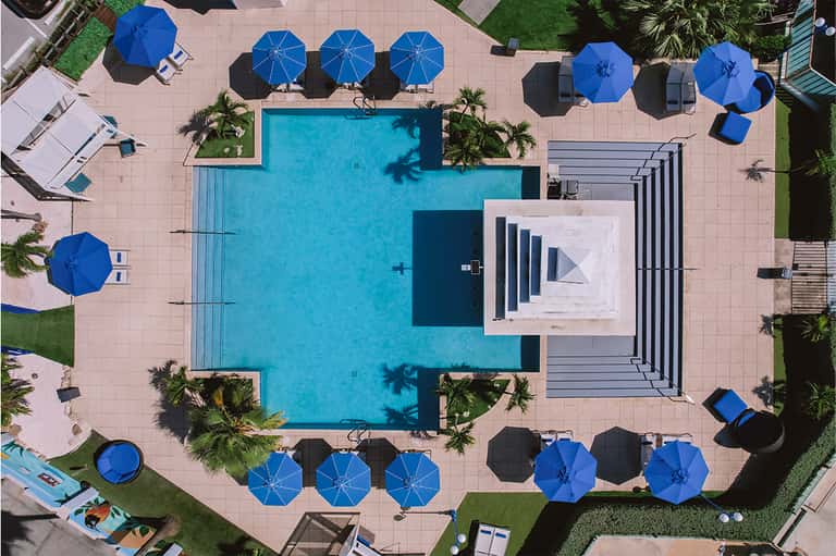 Aerial view of resort pool surrounded by blue umbrellas, palm trees, and manicured lawns