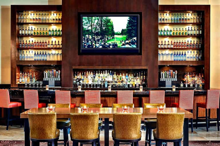 Upscale bar lounge with illuminated bottle displays, dark wood backdrop, and TV screen with gold-accented seating.
