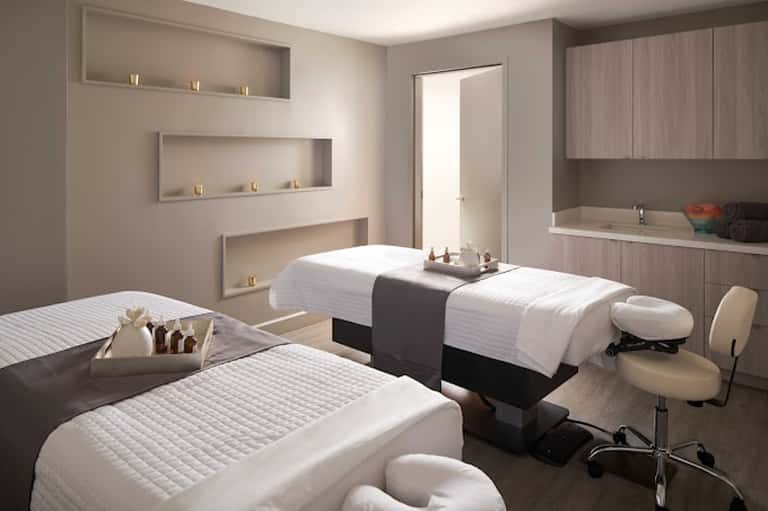 Modern spa treatment room with massage beds, recessed wall shelving, built-in bathtub, and neutral tones